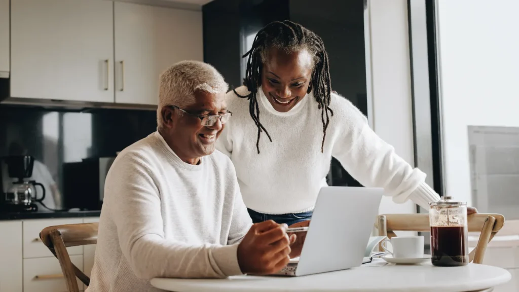 A happy senior couple smiling while reviewing their retirement finance updates on a laptop at a kitchen table, illustrating the positive impact of the 12% average SSNIT Pension Increase for 2025 on Ghanaian retirees.