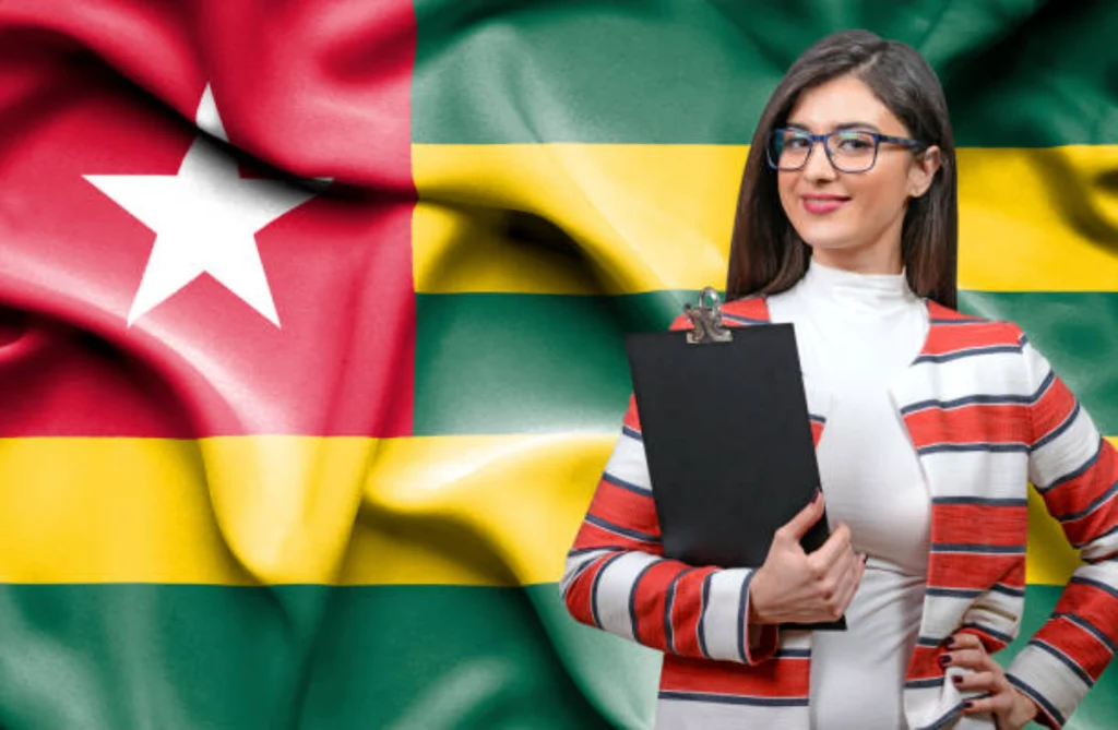 A smiling professional businesswoman wearing glasses and holding a clipboard, standing in front of a waving national flag of Togo, illustrating career opportunities and compliance rules for working in togo as a foreigner.