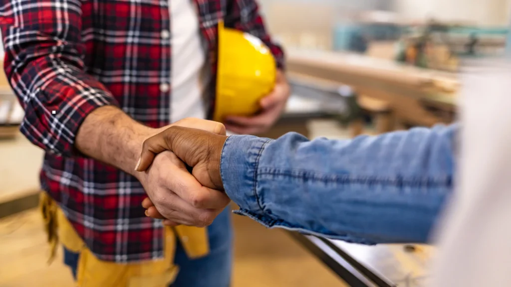A close-up, high-angle view of two technical workers in a construction workshop shaking hands, with one holding a yellow hard hat, representing a smooth operational partnership managed by contractor management services in Ghana